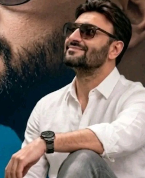 Cristiano Max de camisa branca e óculos escuros sorrindo em frente a um cartaz grande